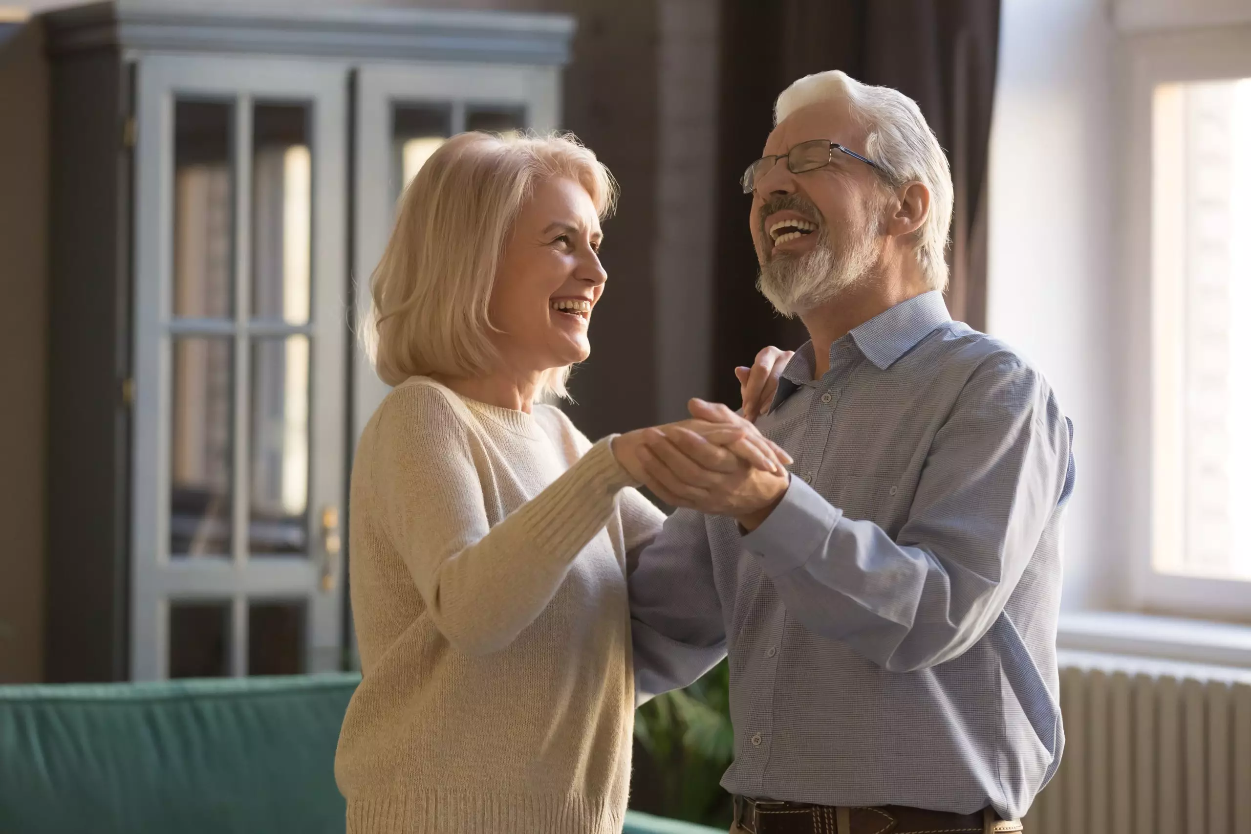 Laughing aged spouses holds hands standing in living room dancing at home